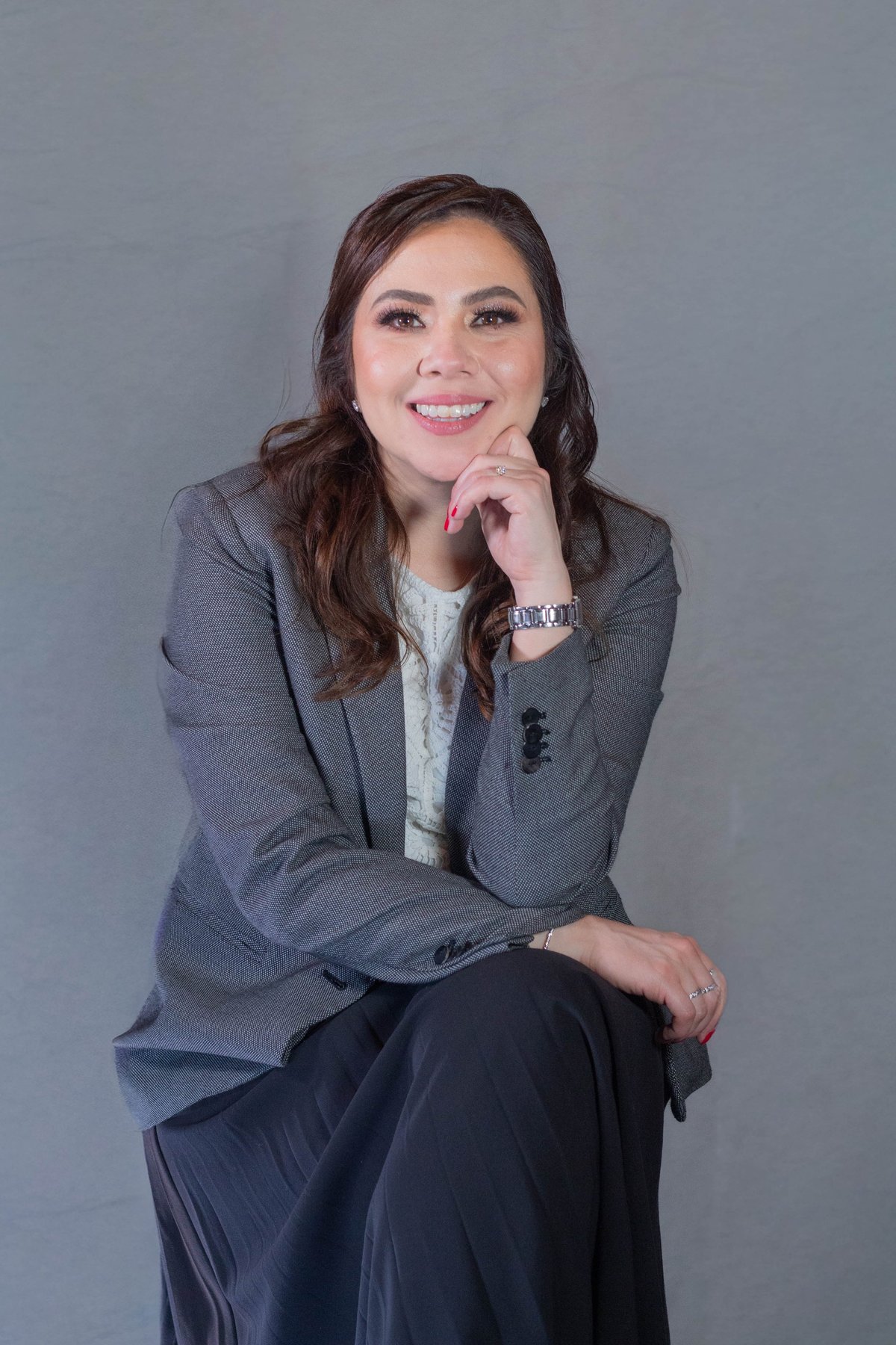 Professional headshot of a smiling woman in a gray blazer and dark pants, sitting with hand near face against gray background