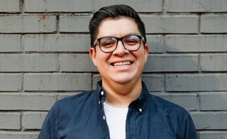 Smiling man wearing glasses and navy button-up shirt against gray brick wall
